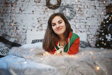 A girl in a New Year's overalls with a festive mood lies on a bed around luminous garlands