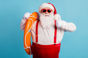 Fototapeta na wymiar Photo of retired old man grey beard hold inflatable circle show thumb-up protect beach ready save life wear red santa x-mas costume suspender sunglass cap isolated blue color background