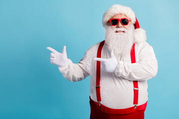 Fototapeta na wymiar Photo of pensioner grandfather grey beard direct finger empty space suggest christmas decoration sale wear red santa costume suspenders spectacles white cap isolated blue color background