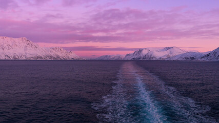 Stunning panorama view from the stern of a cruise ship with the vessel's wake in the arctic sea, snow-covered mountains and a beautiful purple colored sky after sunset in winter time. - obrazy, fototapety, plakaty