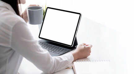 Woman holding mug and digital pen sitting  at the table and using tablet, blank screen for graphic design.
