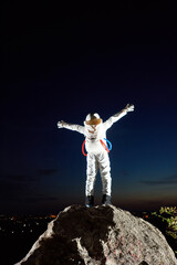 Back view of space traveler standing on top of rocky mountain and spreading hands in sides. Astronaut raising arms while looking at fantastic night sky. Concept of cosmonautics, space travel, freedom