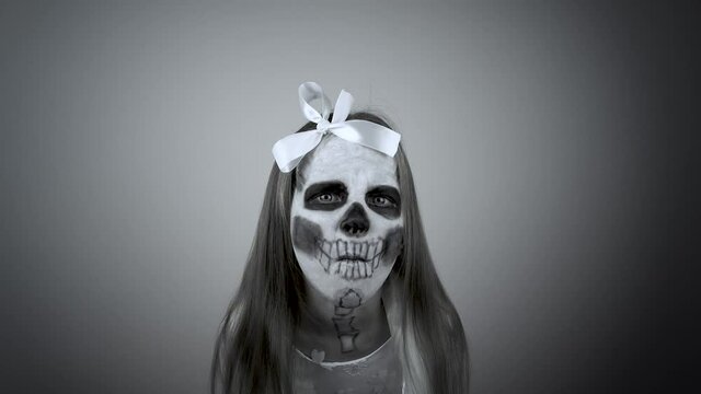 A little girl in a beautiful white dress with a terrible make-up on her face scares. Scary image of a skull on the face. Halloween costume. Face painting.4k
