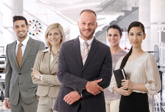 Group of successful business people at bright office, standing, smiling, happy, looking at camera. Suit.