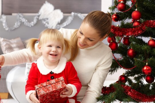 Happy little girl holding christmas present, sitting on mothers' lap by christmas tree.