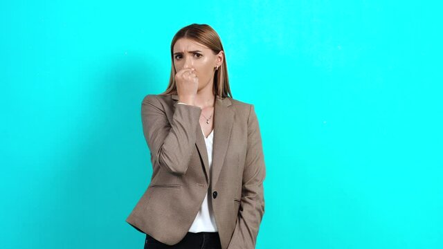 Intelligent young woman, in business style, who closes her nose with her fingers, feeling an unpleasant disgusted smell. Studio image indoors, isolated on blue background