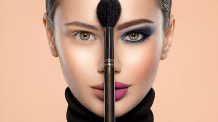 Portrait of a girl with cosmetic brush at face. Woman holds a big makeup brush right in the middle of the face. One half face of a beautiful white woman with  bright makeup and the other is natural.