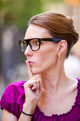 Urban portrait of a dark haired woman with black glasses looking away, with finger on mouth.