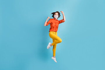 Fototapeta na wymiar Full length photo of crazy lady jump fists rejoicing wear mask orange shirt yellow trousers sneakers isolated blue color background