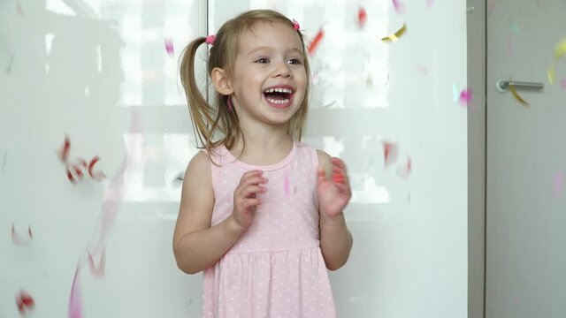 Portrait of little funny girl laughs, smiles and catches multicolored confetti. Kid gets scared and flinches by loud sound of petard. Happy child celebrates birthday.