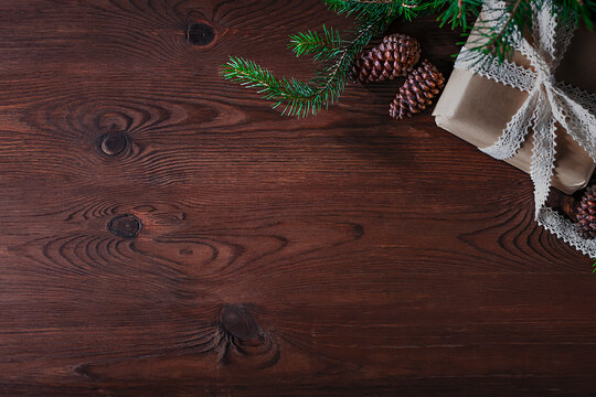 On a wooden, brown background lies a box with a gift, a sprig of a Christmas tree and cones.