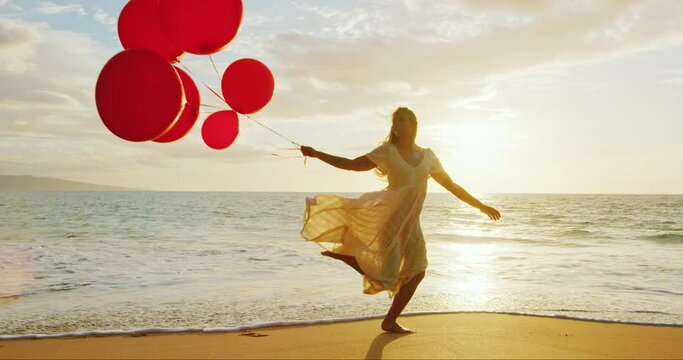 Beautiful happy young woman running down the beach holding red balloons at sunset, slow motion