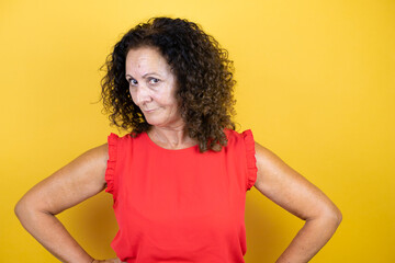 Middle age woman wearing casual shirt standing over isolated yellow background skeptic and nervous, disapproving expression on face with arms in waist