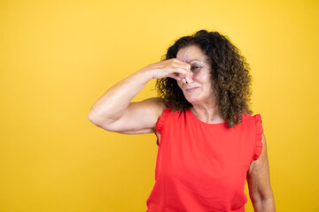 Middle age woman wearing casual shirt standing over isolated yellow background smelling something stinky and disgusting, intolerable smell, holding breath with fingers on nose