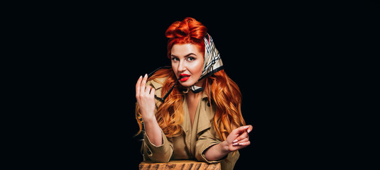 A beautiful red-haired girl on a black background dressed in the style of pin-up with red lipstick holds glasses and look at the camera. Valentine's Day, March 8, International Women's Day