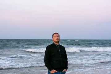 Young Caucasian man stands on the beach in evening. Big waves with white foam at sunset. Selective...