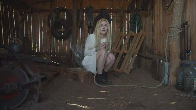 Ruthless criminal kidnapper with bloody axe threatening terrified helpless attractive woman with hands tied up, hidden in corner of dark rustic shed, expressing fear, horror, hopelessness and despair.