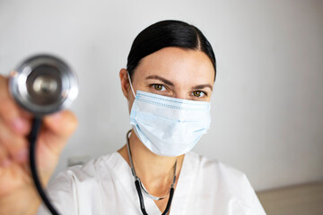 A female doctor in a mask is holding a stethoscope and smiling while looking into the camera.