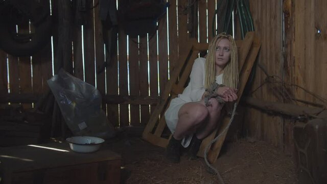 Ruthless kidnapper feeding terrified abducted woman, flinging dirty bowl with food in rustic shed. Scared kidnapped female with hands tied up with rope looking with hunger on meal served by criminal.