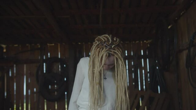 Terrible young attractive dead ghost woman with dreadlocks and pale face dressed in white dress standing in dark rustic shed, looking with furious eyestare, expressing fear, horror and anger.