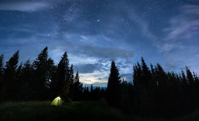 Panoramic view of night starry sky with illuminated tourist tent in forest with coniferous trees under magical blue sky with stars. Concept of travelling, hiking and night camping.