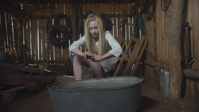 Spooky ghost drowned woman with pale face and dreadlocks wearing white dress, washing tattered clothes in rusty dirty trough in dark shed, being restless and lost after death.