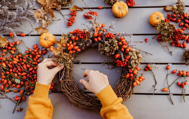 Girl making floral autumn door wreath using colorful rosehip berries, rowan, dry flowers and pumpkins. Fall flower decoration workshop, florist at work. - Powered by Adobe