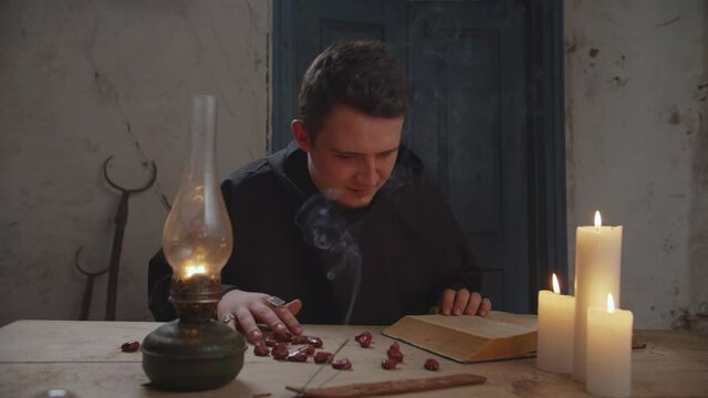 Mysterious wizard in black robe casting rune stones on rustic table and using ceromancy method of divination, reading candle's melted and hardened wax and interpreting signs into prediction.
