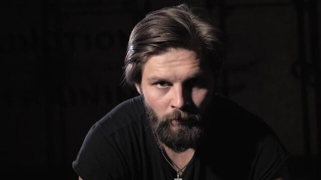 Close up view of a young athlete with a beard sits on a tire in a dark gym, raises his head and looks at the camera. Sport motivation.