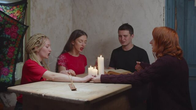 Diverse mystical people with eyes closed holding hands, sitting around table with candle lights and saying evocation hexes, frightened by paranormal activity during summoning spirits in rustic house.