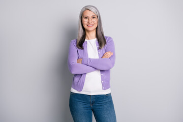 Photo of charming pretty old lady crossed hands self-assured toothy beaming smile wear white shirt purple cardigan glasses denim jeans isolated grey color background