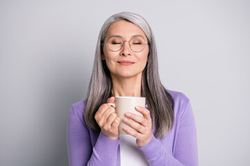 Photo portrait of woman holding cup of tea or coffee with two hands enjoying smell and taste with eyes closed wearing casual violet shirt isolated on grey colored background