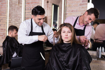 Two skilled men hairdressers making hairstyle for young female client in hair studio