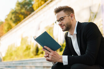 Handsome focused businessman reading book while sitting on bench