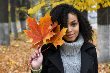 Portrait of a beautiful young African American woman with orange maple leaves in autumn park.