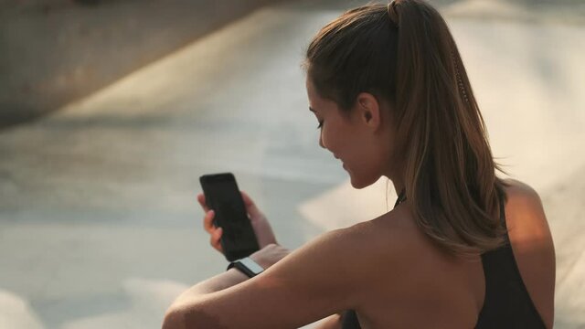 A back view of a young woman is using her fitness tracker sitting at a sports ground outside