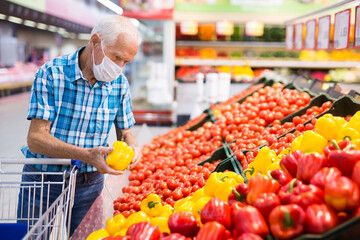 elderly caucasian man in mask with covid protection choosing tomatoes in vegetable section of supermarket