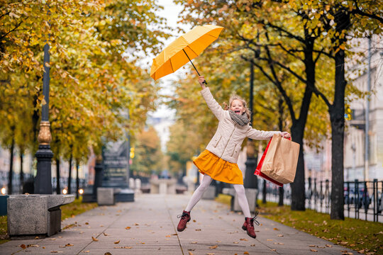 Happy girl in a jump on the autumn city street with bright yellow umbrella and shopping bags.