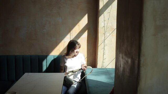 A modest young girl in a white T-shirt and jeans sits at a table by the window in a cozy city cafe. Close-up