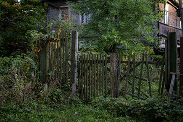 Naklejka premium Old rickety wooden fence made in front of village house among autumn trees with foliage and green dense grass. Russian abandoned building