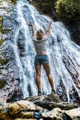 Young caucasian slender woman tourist in casual clothes and sneakers stands on a rock and look at waterfall 