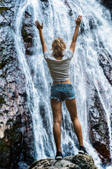 Young caucasian slender woman tourist in casual clothes and sneakers stands on a rock and look at waterfall 