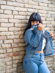 Young woman cover her face with her denim jacket in front a brick wall. Long dark hair natural girl portrait. Playful and cheerful female.