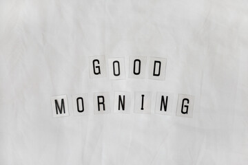 Fototapeta na wymiar Inscription Good morning, composed of black letters on transparent plastic bases on white rumpled sheets. Top view, flat lay. Horizontal. Concept of rest, awakening, sleep. Minimal style