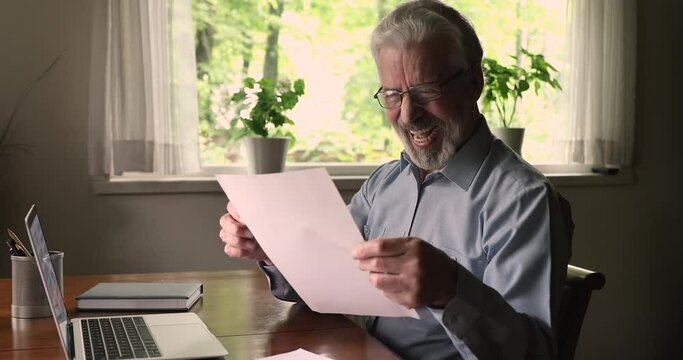 Happy aged man sit at table hold sheet read letter feels excited by super news. Euphoric senior male celebrate bank loan approve or last mortgage payment, satisfied client received great offer concept