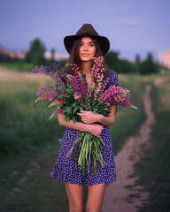 Girl with lupines in a lilac dress and hat on the path