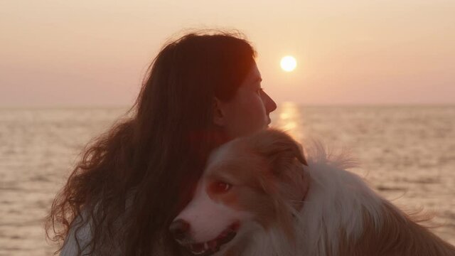 Close up portrait Lonely brunette woman kissing at the beach with her dog at sunset on the background of sun going down. Young girl hugging her border collie dog. Best friends, concept of friendship