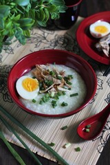 Breakfast concept with chinese style, a bowl of chicken rice porridge topping with chicken slices, boiled egg, and spring onion. Oriental set ware on bamboo place matt. 