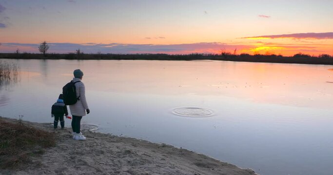 Mother with Child Throw Sand in Water near Wooden Fishing Pier on River Lake Pond Shore. Scenic Picturesque Sunset Cloudscape Reflection Beautiful Evening Landscape