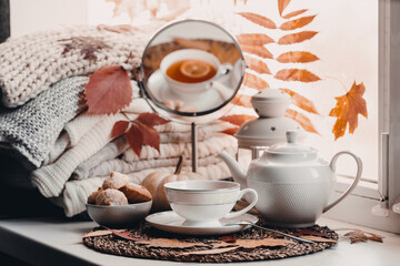A cup of hot tea with lemon in an early autumn morning by the window. A stack of sweaters, yellow leaves, pumpkin, candles. Decor. Autumn concept of warm drink on a cold day.